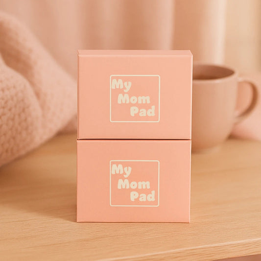 Deux boîtes roses superposées "My Mom Pad", posées sur une table en bois, dans un décor doux et chaleureux. Produits d’hygiène post-partum.