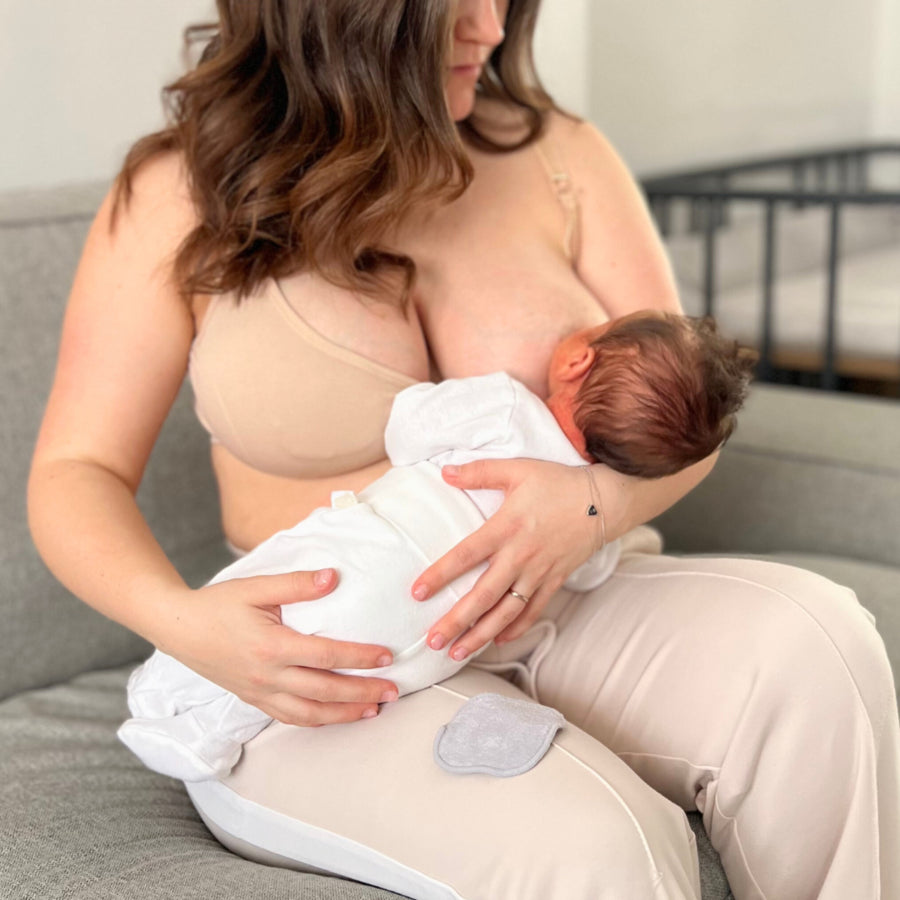 Maman en brassière d’allaitement beige nourrissant son bébé en position madone sur un canapé utilisant un coussinet lavable sous sa brassière, avec un coussinet lavable posé à côté d’elle.