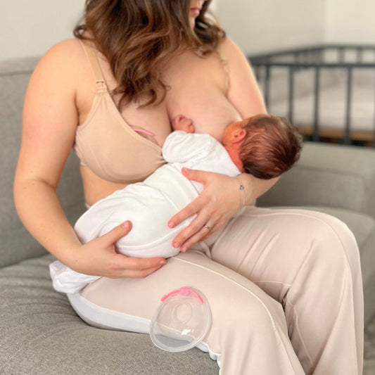 Maman en brassière d’allaitement beige nourrissant son bébé en position madone sur un canapé utilisant un recueil-lait sous sa brassière, avec un recueil-lait posé à côté d’elle.