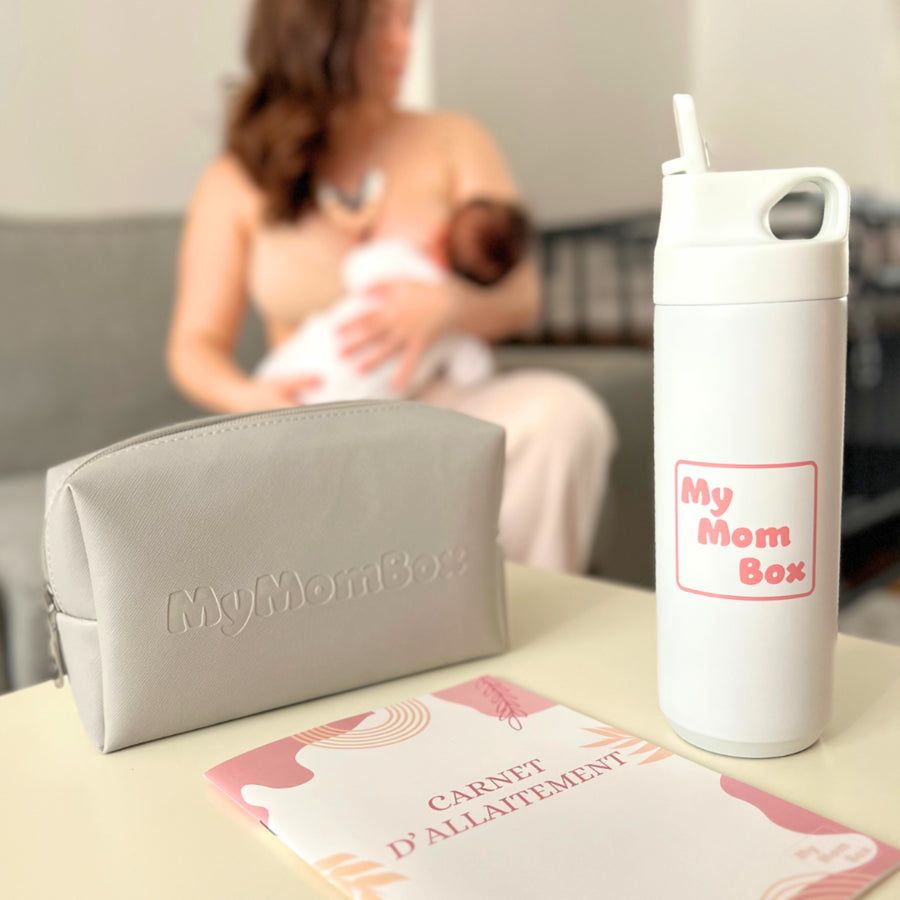 Gourde blanche MyMomBox, trousse de rangement et carnet d’allaitement posés sur une table, avec en arrière-plan une maman allaitant son bébé sur un canapé.