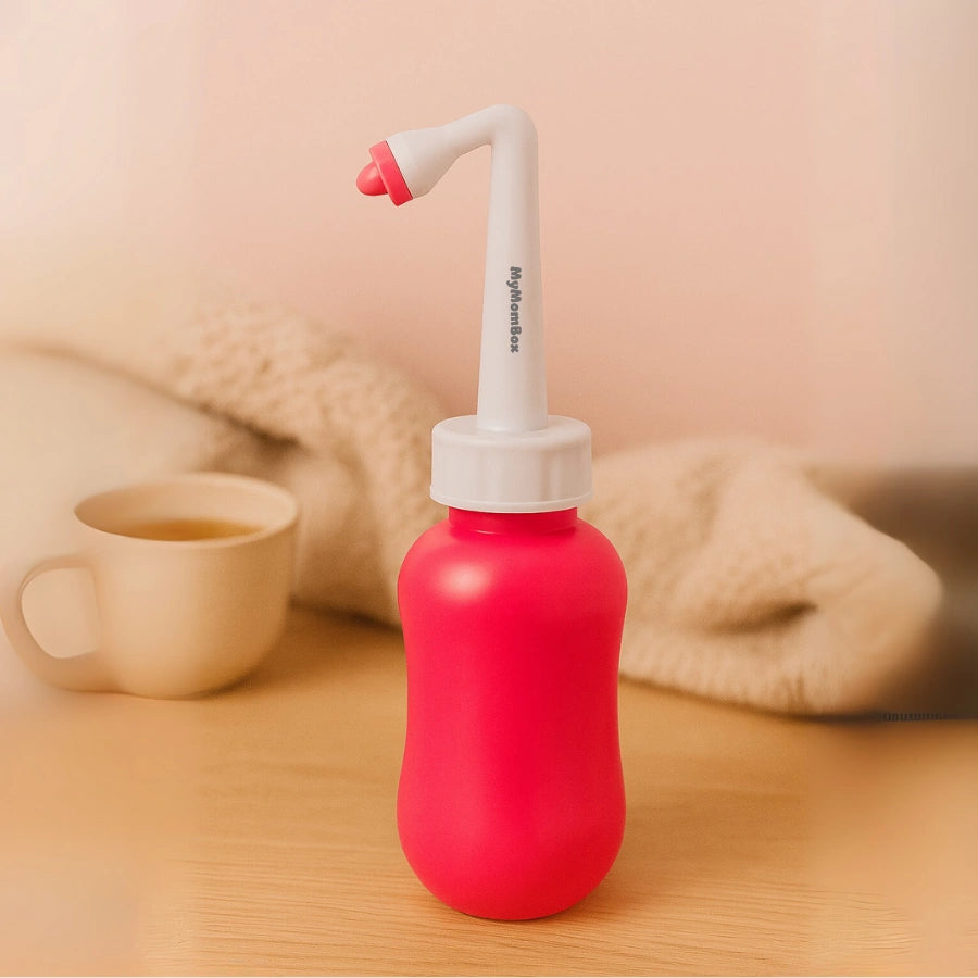 Bidet portable (bouteille de lavage intime) post-partum rose et blanche avec embout ergonomique, posée sur une table en bois, pour soulager les mamans après l'accouchement. Marquée "MyMomBox".