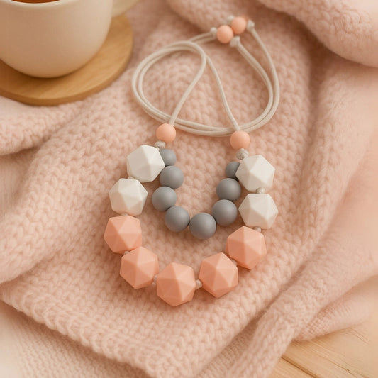Collier d’allaitement ou sensoriel en perles de silicone pastel (rose, gris, blanc), posé sur une couverture tricotée. Utile pour occuper bébé pendant l’allaitement.