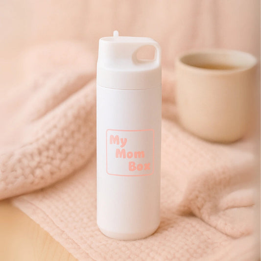 Gourde en acier inoxydable 550ml blanche avec logo rose "My Mom Box", posée sur une couverture rose. Conçue pour garder l’eau chaude ou froide, pratique pour les mamans actives.